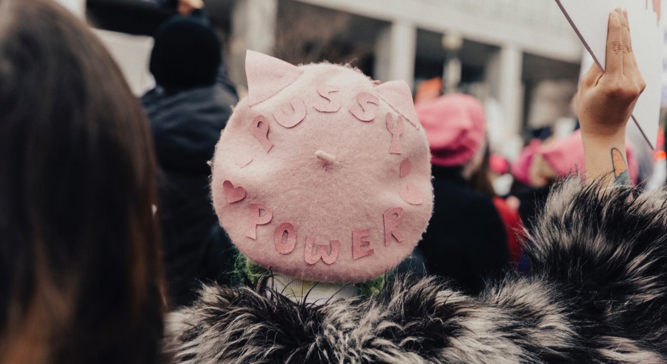 selective focus photography of person wearing pink fleece Pussy Power cap raising right hand