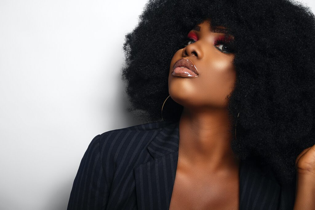 Photo for ENTITY Mag of a woman in full makeup with an afro looking confident and wearing a striped blazer on a gray background.