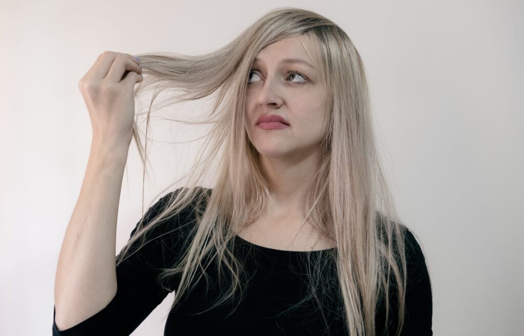 Photo of a woman for ENTITY Mag holding a lock of hair and looking at it in disgust on a gray background. Hairstyles quotes. 